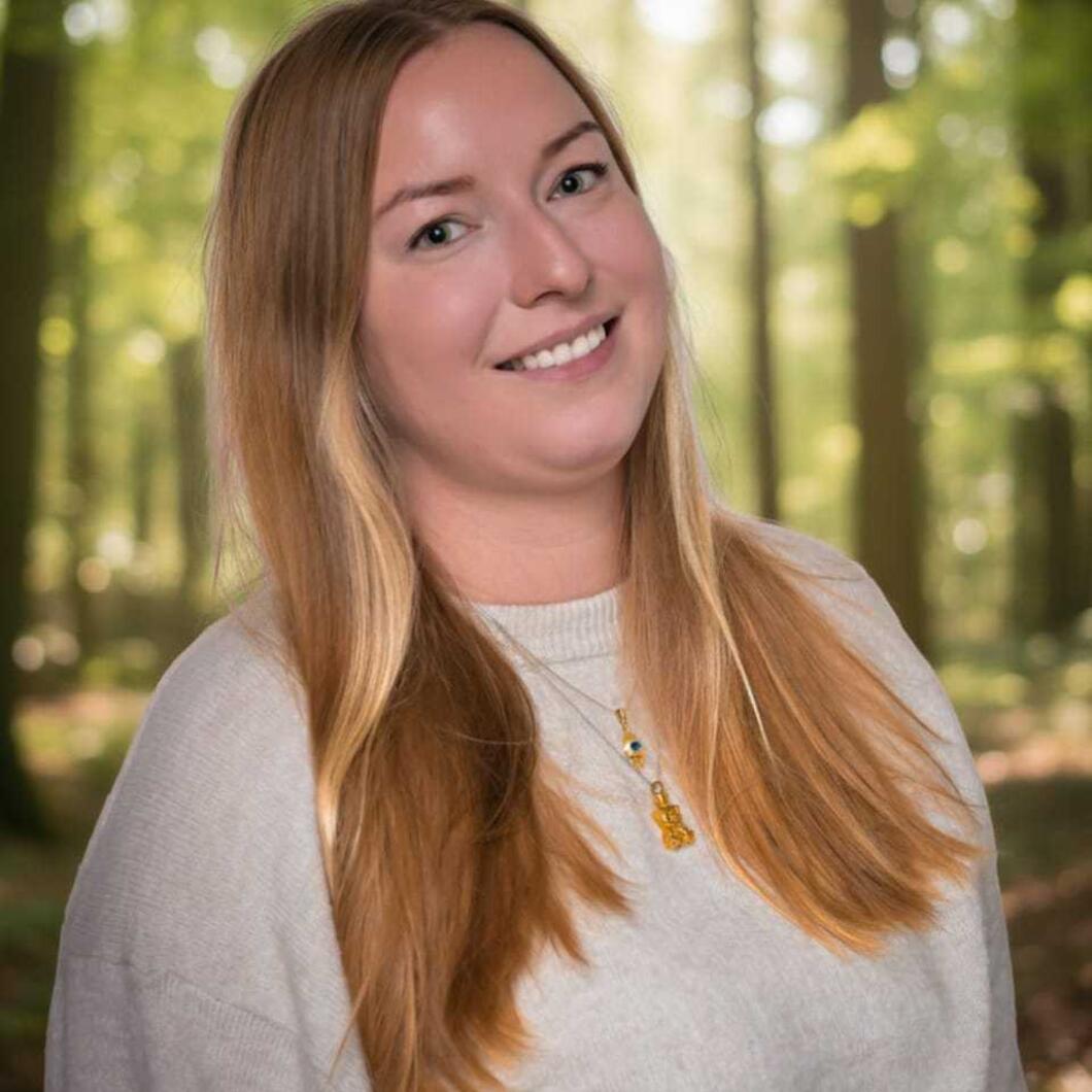 We zien een vrouw met lang blond haar en op de achtergrond bomen waar de zon tussendoor schijn.t 