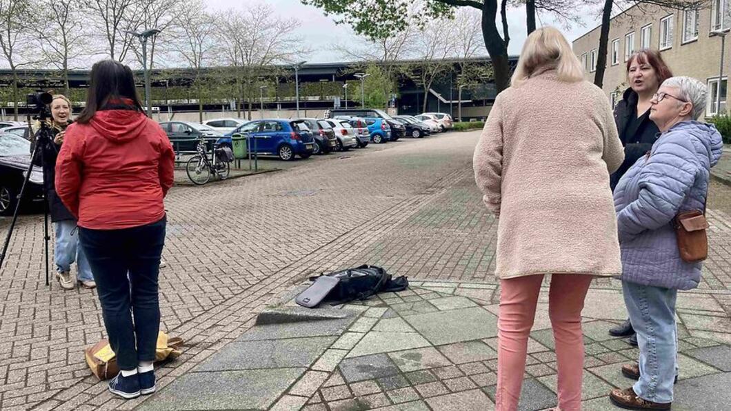 Groepje van 3 dames, op de rug gezien, rechts in beeld. Links zien we een vrouw met rode jas op de rug, daarvoor een statief met camera en een dame met microfoon die haar interviewt.