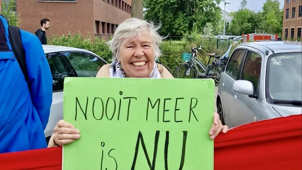 Vrouw achter rode lijn houdt groen protestbord vast met tekst ‘Nooit meer is NU’. 