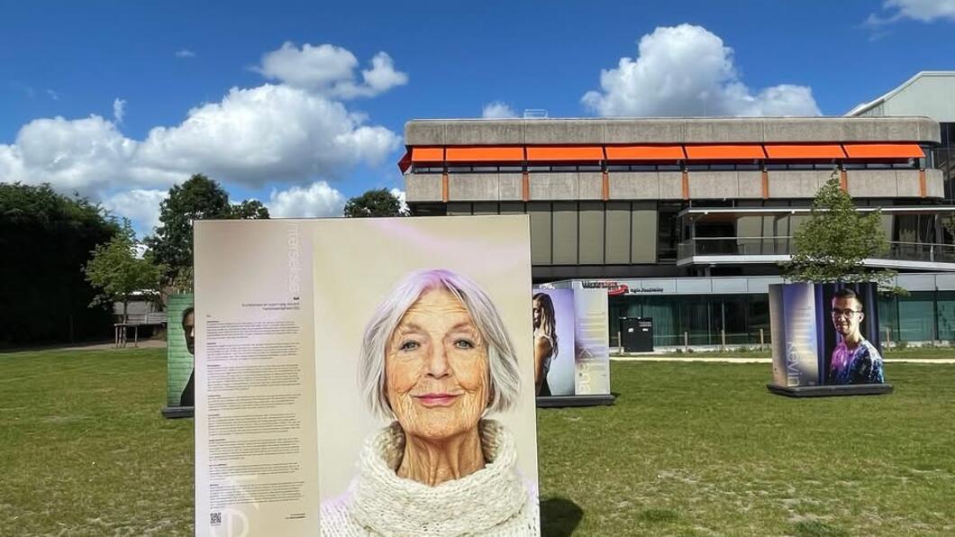 Een oudere vrouw met halflang grijs haar kijkt de camera in, zij staat op een bord dat onderdeel is van de buitenexpositie ‘1 op 90’