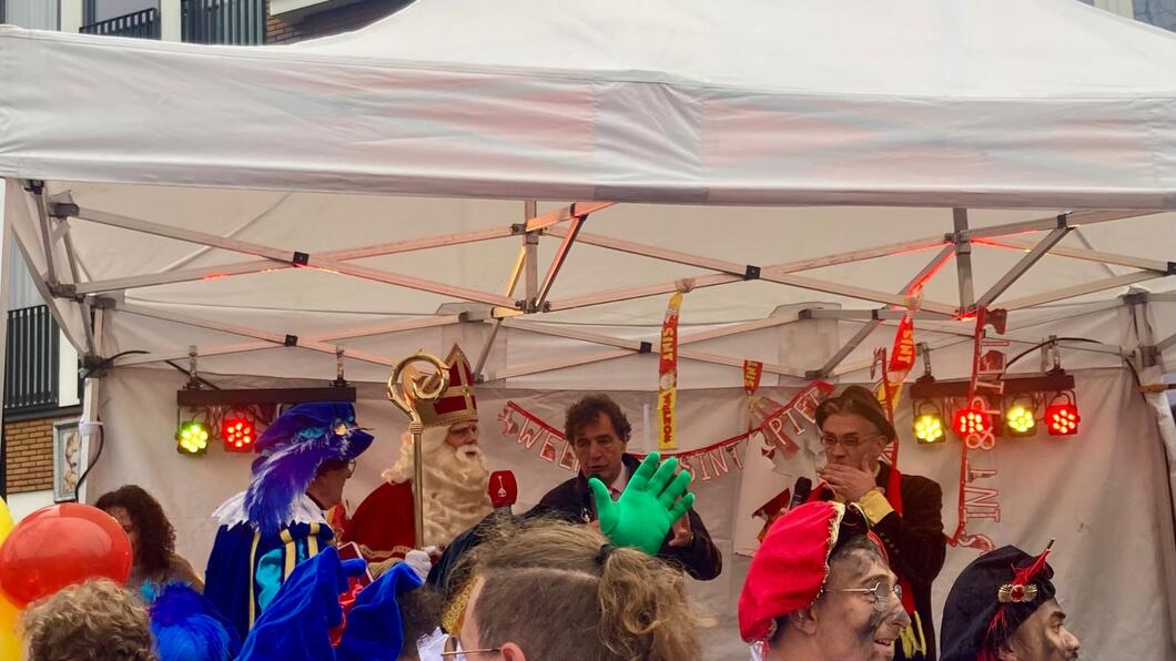Groep mensen (ouders en kinderen) op de rug gezien en je kijkt tegen een witte tent met daarin de burgemeester en Sinterklaas. Ervoor enkele roetveeg pieten.