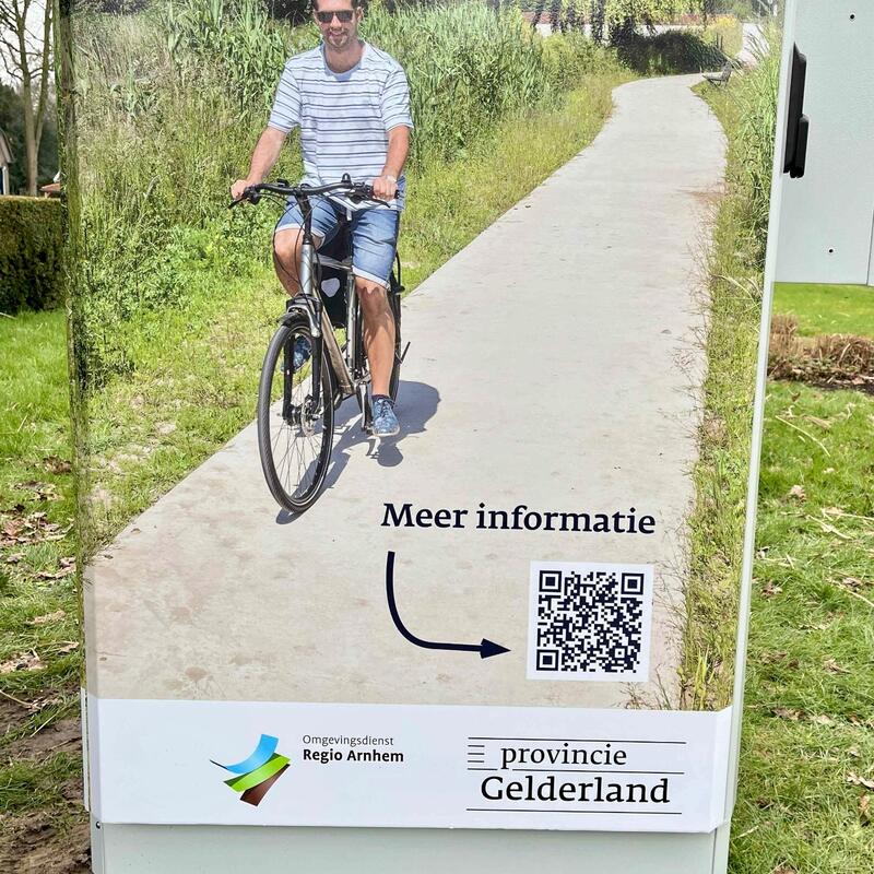 Foto van bestickering van het luchtmeetstation - daarop een QR-code en een man op een fiets tussen groene omgeving. 