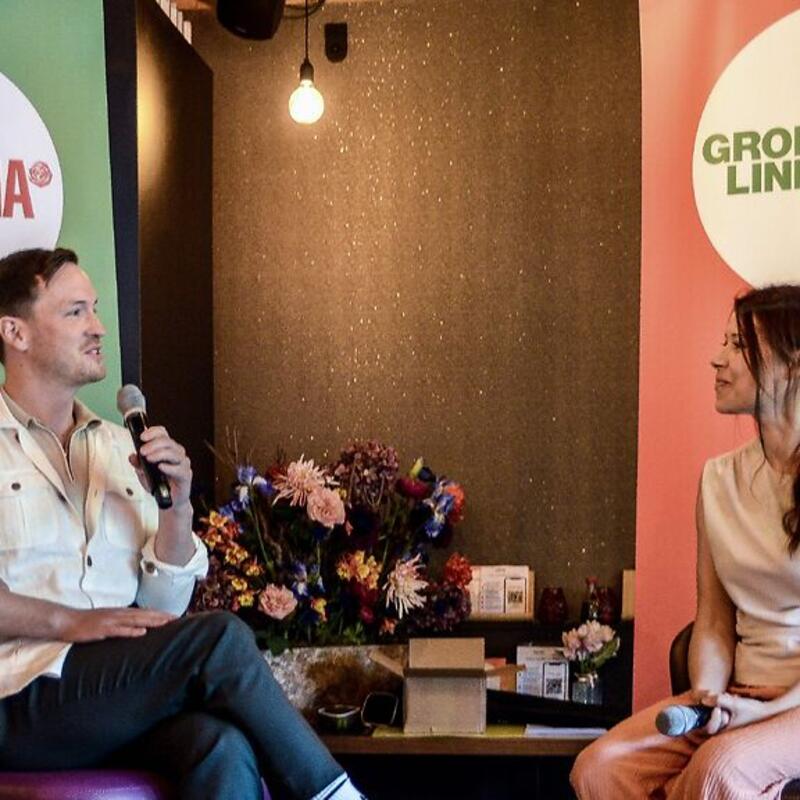 Op een podium zit voor een groene banner met ‘GroenLinks PvdA Ede’ erop zit links raadslid Robin Bongers-Karmaoui rechts zit voor een rode banner Tweede Kamerlid Lisa Westerveld.