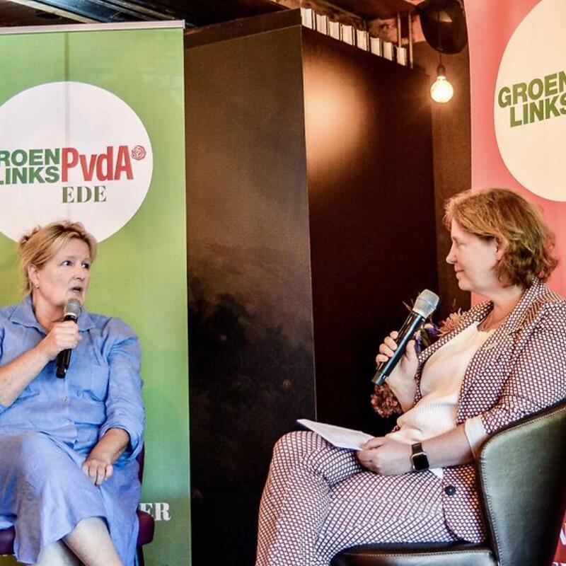 Voor een groene banner met ‘GroenLinks-PvdA Ede’ zit links Mariette Patijn Tweede Kamerlid en rechts voor een rode banner Karin Bijl PvdA-lid en in Ede wethouder.