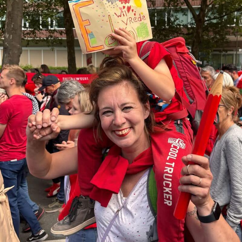 Vrouw met een groot rood potlood in de hand en op haar nek een kind dat z’n gezicht verbergt achter een A4 waarop staat: “Ede(n) wil vrede!”