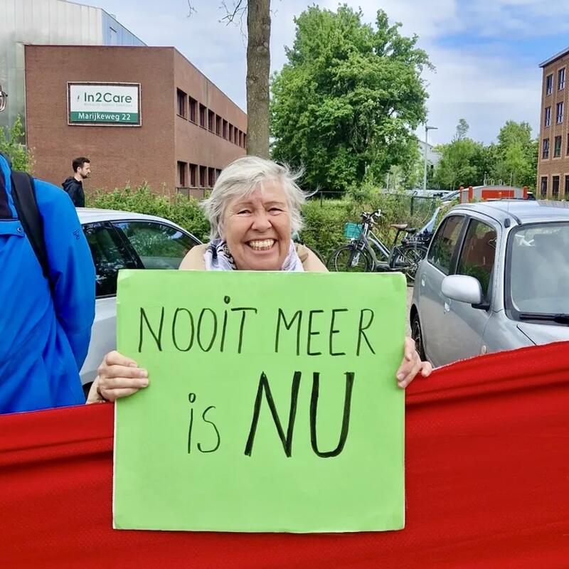 Vrouw achter rode lijn houdt groen protestbord vast met tekst ‘Nooit meer is NU’. 