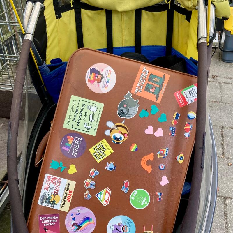 Bruine koffer vult een fietskar, de koffer is bestickerd met een verzameling stickers. 