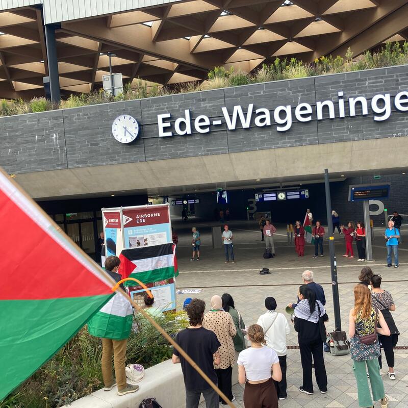 Foto op station Ede-Wageningen waar de combinatie van woonplaatsen zichtbaar is en links in beeld een deel van de Palestijnse vlag in beeld.