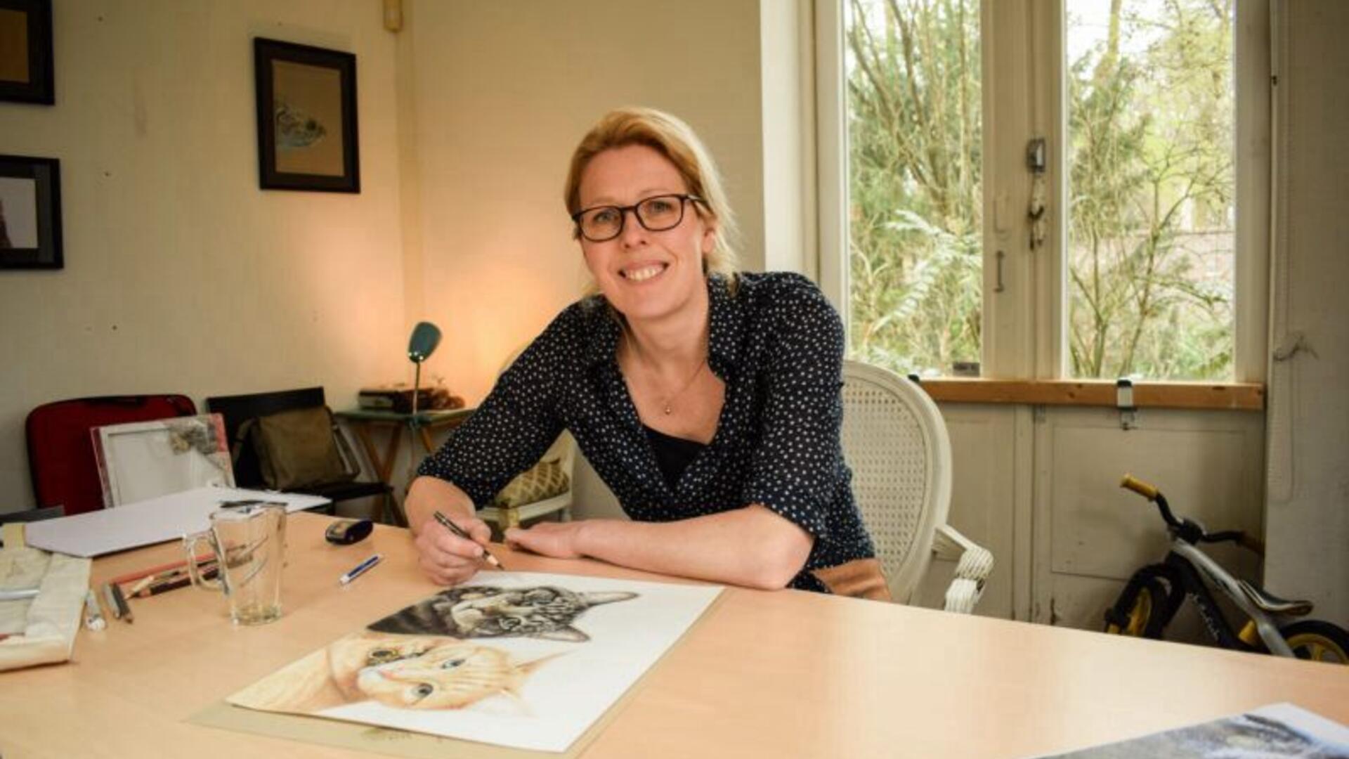 Vrouw wit met rood haar zit aan een tafel. Op de tafel ligt een potloodtekening van 2 katten.