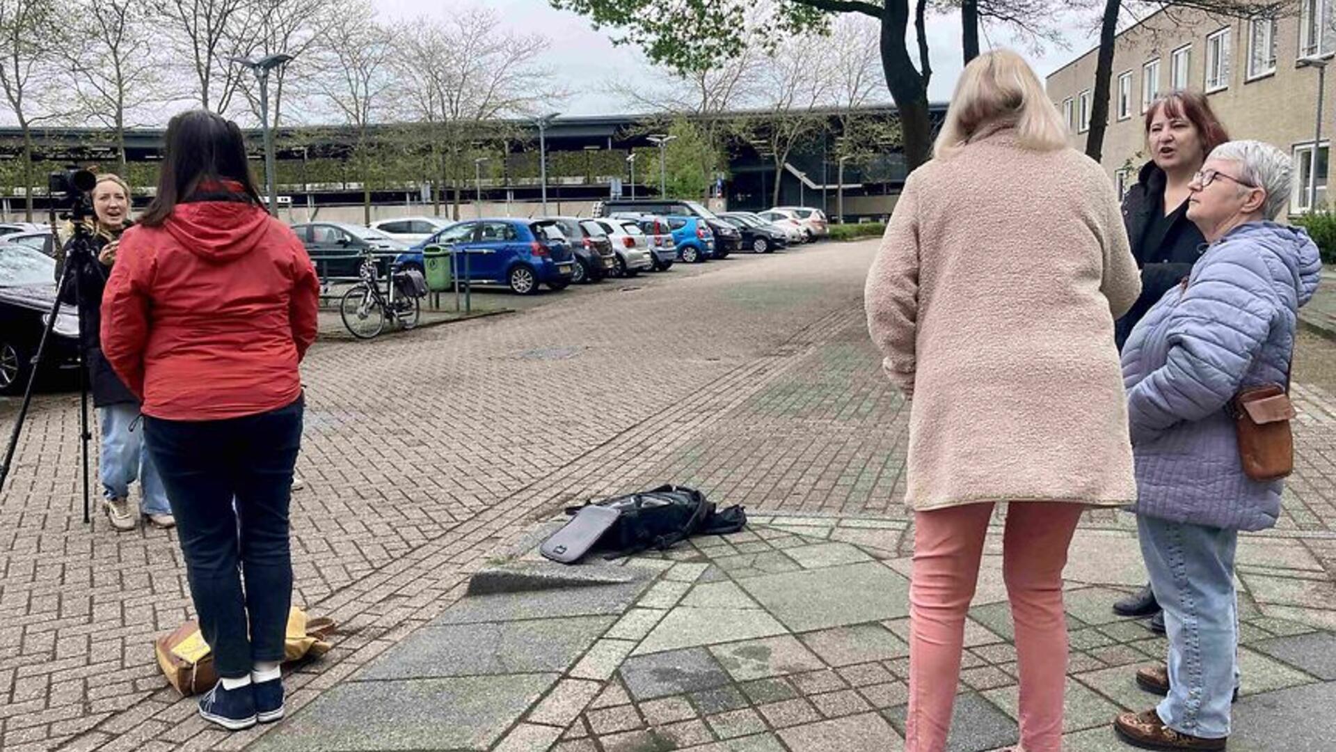 Groepje van 3 dames, op de rug gezien, rechts in beeld. Links zien we een vrouw met rode jas op de rug, daarvoor een statief met camera en een dame met microfoon die haar interviewt.