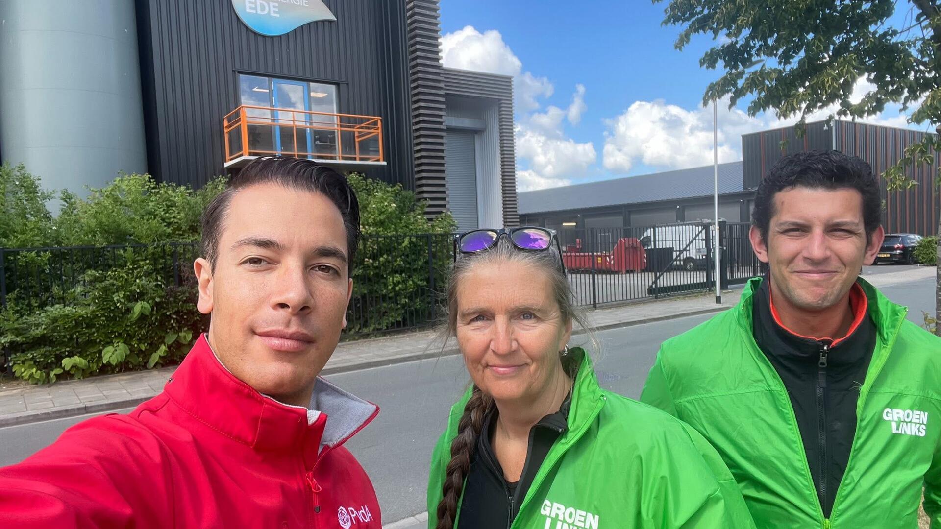 Een persoon in rode jas en twee in groene jassen staan (leden van GroenLinks-PvdA) voor hoog gebouw, de biomassacentrale.