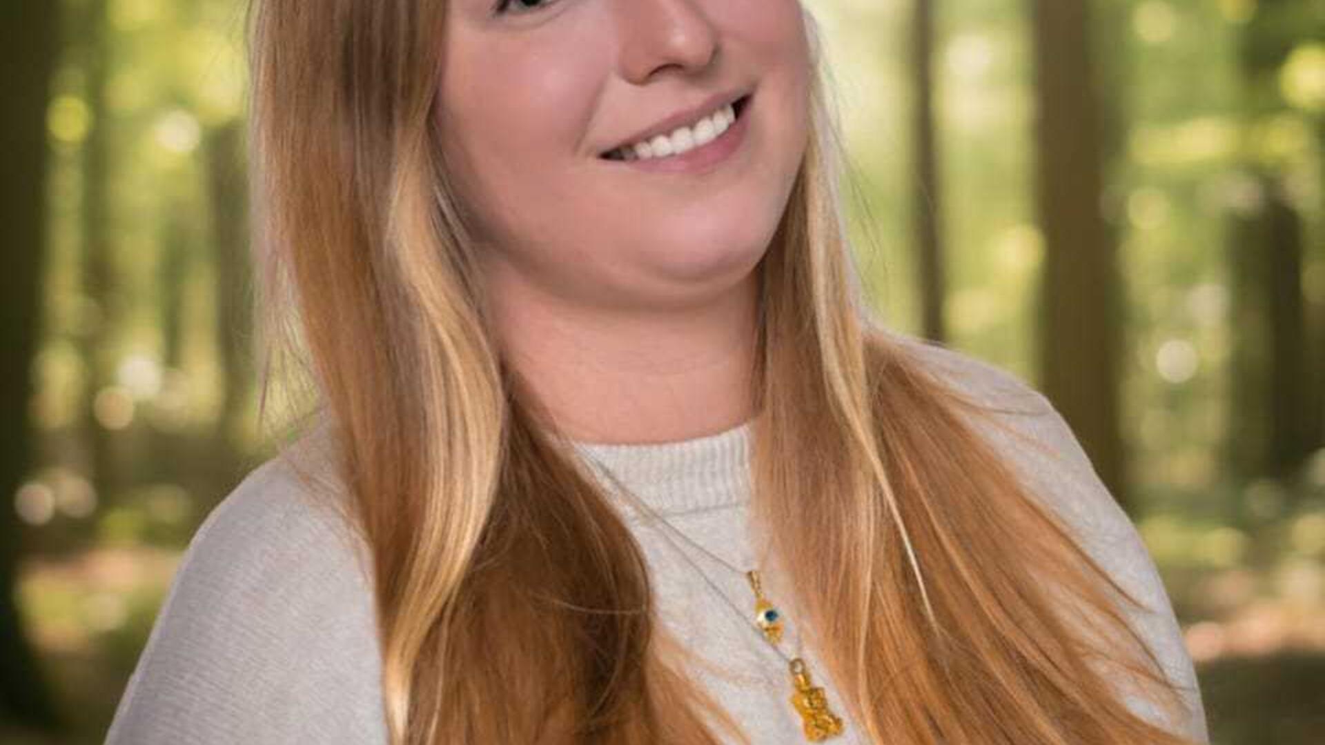 We zien een vrouw met lang blond haar en op de achtergrond bomen waar de zon tussendoor schijn.t 